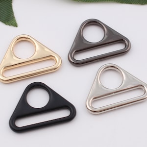 Pode incluir: Quatro anéis de metal em forma de triângulo em ouro, prata, preto e cinza escuro. Cada anel tem um pequeno círculo na parte superior e uma abertura retangular na parte inferior.