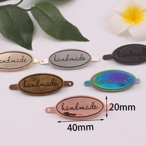 May include: Five oval-shaped metal tags with the word "handmade" engraved on them. The tags are in different colors: gold, silver, black, iridescent, and rose gold. The tags are 40mm long and 20mm wide.