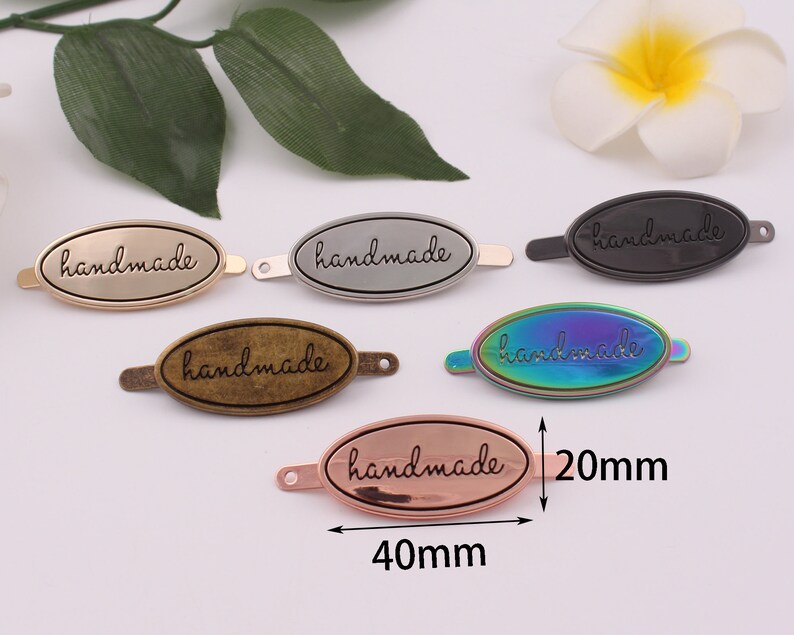 May include: Five oval-shaped metal tags with the word "handmade" engraved on them. The tags are in different colors: gold, silver, black, iridescent, and rose gold. The tags are 40mm long and 20mm wide.