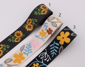May include: Three rolls of black, white, and black fabric ribbon with floral patterns. The black ribbon has a sunflower pattern, the white ribbon has a pink, blue, and brown floral pattern, and the black ribbon has a yellow flower and leaf pattern.