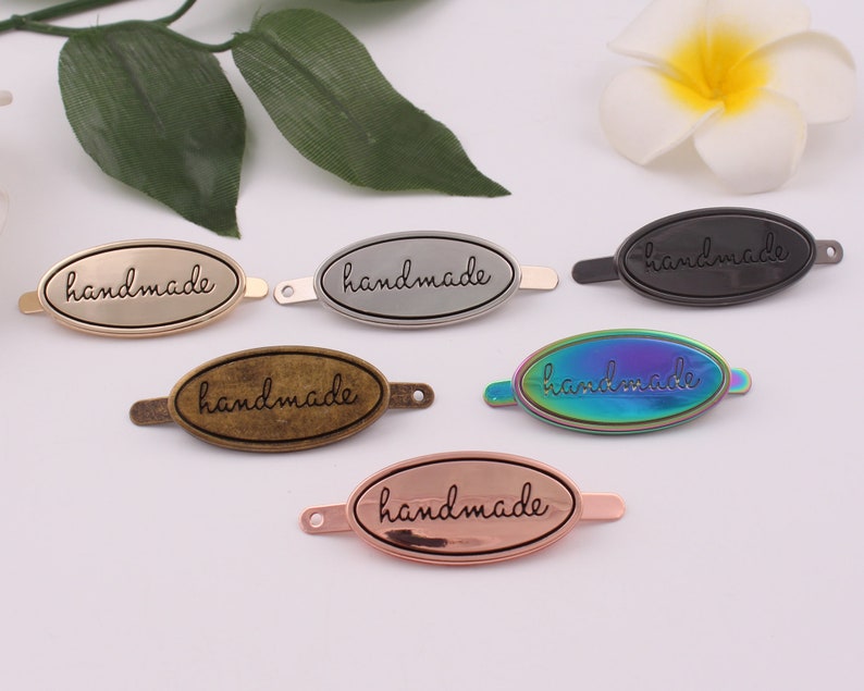 May include: Six oval-shaped metal tags with the word "handmade" engraved on them. The tags are in different colors: gold, silver, black, rainbow, and rose gold.