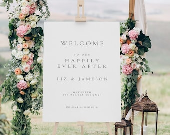 May include: White wedding welcome sign with the text "Welcome to our Happily Ever After Liz & Jameson May Fifth two thousand twentyfour Columbia, Georgia". The sign is propped up on an easel in front of a floral archway.