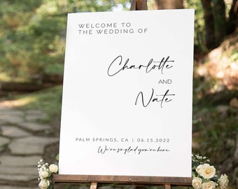 Op de afbeelding: Een wit welkomsbord voor een bruiloft met zwarte tekst. Het bord luidt "Welkom op de bruiloft van Charlotte en Nate, Palm Springs, CA | 06.15.2022, We zijn zo blij dat je er bent."