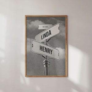 May include: Black and white framed print featuring street signs with the names "LINDA" and "HENRY". A smaller sign reads "EST. 2023". The background shows a cloudy sky. The print is in a wooden frame.