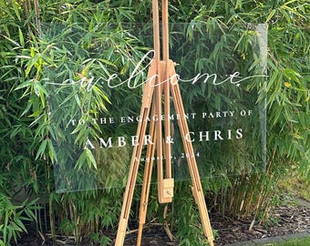 May include: Clear acrylic welcome sign with white calligraphy lettering on a wooden easel. The sign reads "Welcome to the engagement party of Amber & Chris August 6, 2024".