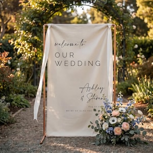 May include: A cream-colored fabric wedding welcome sign hanging from a copper-toned metal frame with white ribbons. The sign features black text reading: welcome to OUR WEDDING, Ashley & Steven, Aug 20 202X, WE'RE SO GLAD YOU'RE HERE.