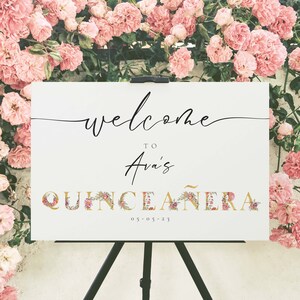 May include: White welcome sign with black calligraphy that reads "Welcome to Ava's Quincea&ntilde;era 05-05-23". The letters are decorated with gold and pink floral accents.