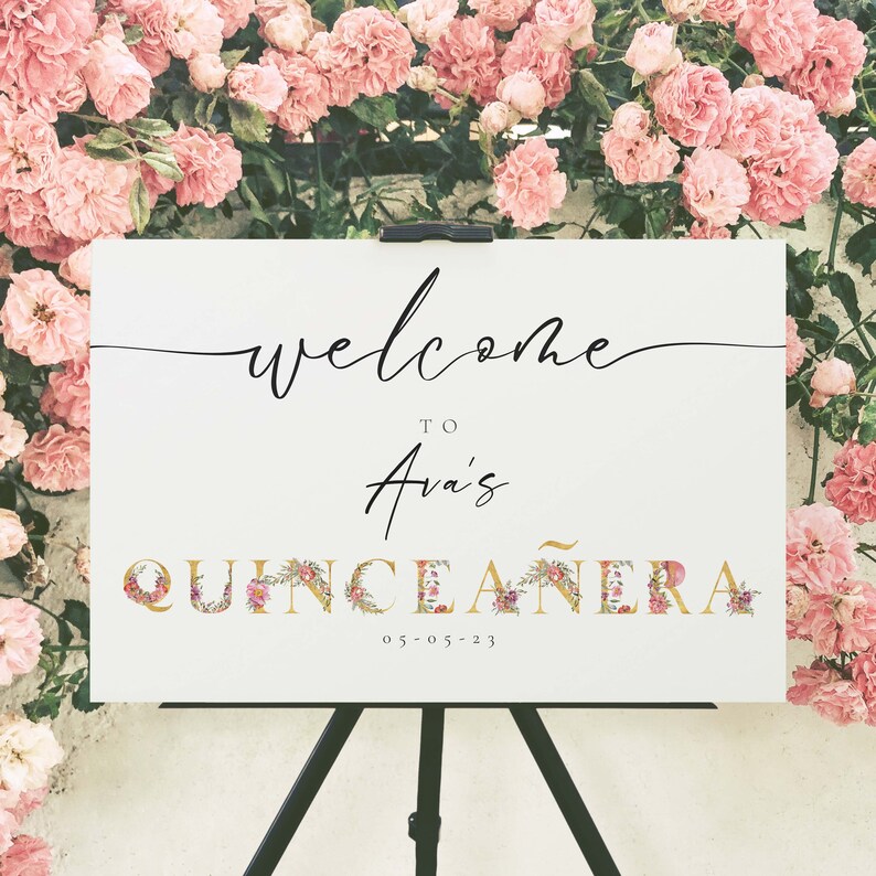 May include: White welcome sign with black calligraphy that reads "Welcome to Ava's Quincea&ntilde;era 05-05-23". The letters are decorated with gold and pink floral accents.