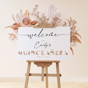 May include: Welcome to Emily's Quincea&ntilde;era sign with floral accents on a wooden easel. The sign is white with black text and a gold and floral design for the word "Quincea&ntilde;era". The date is August 15, 2022.