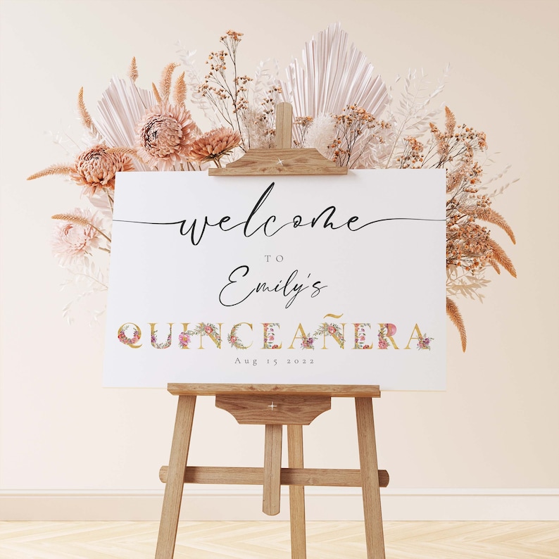May include: Welcome to Emily's Quincea&ntilde;era sign with floral accents on a wooden easel. The sign is white with black text and a gold and floral design for the word "Quincea&ntilde;era". The date is August 15, 2022.