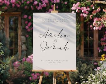 May include: White wedding welcome sign with black calligraphy text that reads "We're so happy you're here! Amelia & Jonah Welcome to our wedding July 19, 2025"