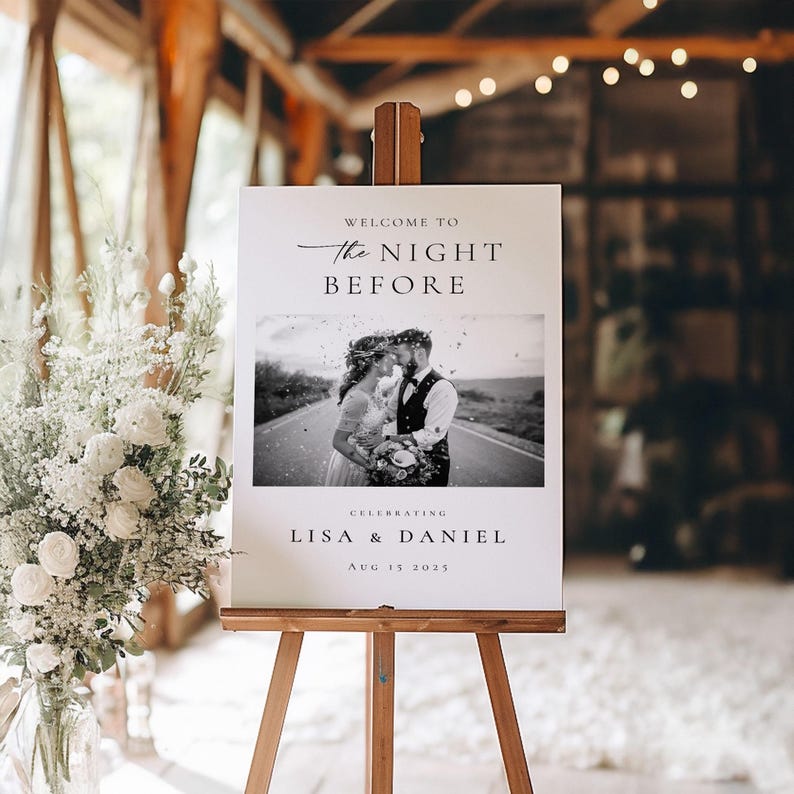 Op de afbeelding: Een welkomstbord voor een bruiloft op een houten ezel. Het bord leest "Welcome to The Night Before" boven een zwart-wit foto van een stel. Daaronder staat "Lisa & Daniel" en de datum "15 aug 2025".