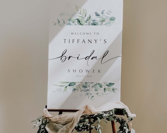 May include: A white sign on a wooden easel with the text "Welcome to Tiffany's Bridal Shower Oct 13, 2024". The sign is decorated with green and gray watercolor eucalyptus leaves. A beige fabric and greenery are draped at the base.