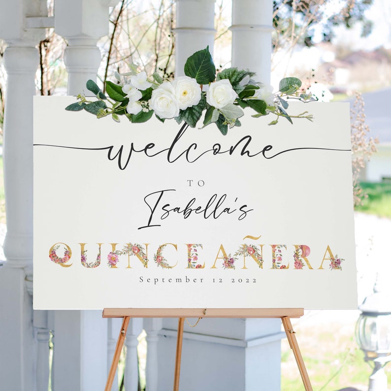 May include: A white welcome sign with floral accents and gold lettering. The sign reads "Welcome to Isabella's Quincea&ntilde;era September 12, 2022".