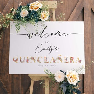 May include: White welcome sign with gold lettering for a Quincea&ntilde;era. The sign says "Welcome to Emily's Quincea&ntilde;era Aug 15 2022". The letters are decorated with pink and purple flowers.