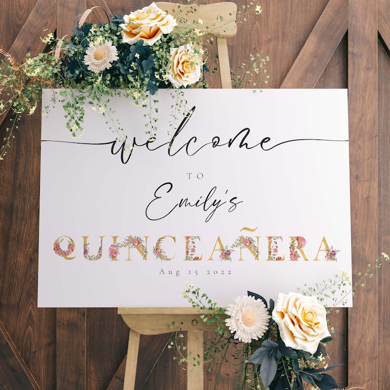 May include: White welcome sign with gold lettering for a Quincea&ntilde;era. The sign says "Welcome to Emily's Quincea&ntilde;era Aug 15 2022". The letters are decorated with pink and purple flowers.
