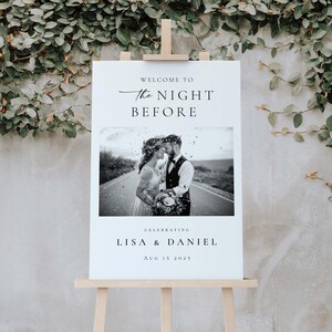 Op de afbeelding: Een wit welkomstbord voor een bruiloft op een houten ezel. Het bord toont de tekst "WELCOME TO THE NIGHT BEFORE" boven een zwart-wit foto van een stel. Onder de foto staat "CELEBRATING LISA & DANIEL AUG 15 2025".