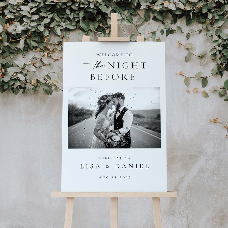 Op de afbeelding: Een wit welkomstbord voor een bruiloft op een houten ezel. Het bord toont de tekst "WELCOME TO THE NIGHT BEFORE" boven een zwart-wit foto van een stel. Onder de foto staat "CELEBRATING LISA & DANIEL AUG 15 2025".