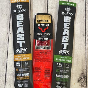 Peut inclure: Trois b&acirc;tonnets de viande emball&eacute;s individuellement. L'emballage noir et or indique "Honey BBQ" et "Beast Stick". L'emballage rouge indique "Original 11g Protein Beef Steak". L'emballage noir et vert indique "Jalape&ntilde;o" et "Beast Stick".