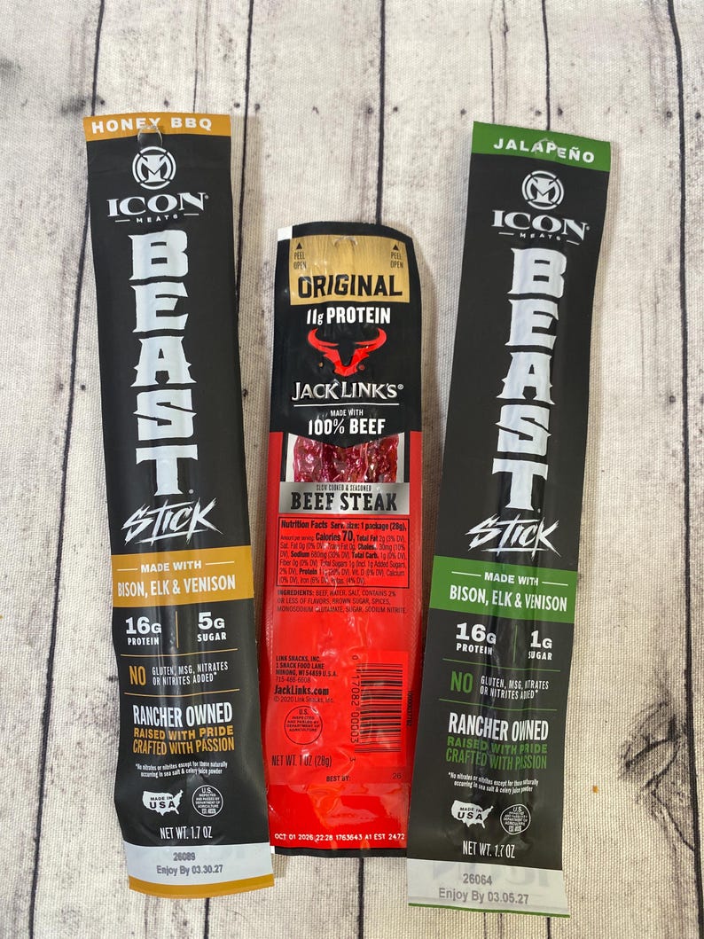 Peut inclure: Trois b&acirc;tonnets de viande emball&eacute;s individuellement. L'emballage noir et or indique "Honey BBQ" et "Beast Stick". L'emballage rouge indique "Original 11g Protein Beef Steak". L'emballage noir et vert indique "Jalape&ntilde;o" et "Beast Stick".