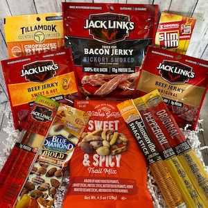 Peut inclure: Un assortiment d'en-cas emball&eacute;s, dont du jerky de b&oelig;uf, du jerky de bacon, du m&eacute;lange montagnard et des b&acirc;tonnets de collation. Les marques incluent Jack Link's, Slim Jim, Tillamook, Blue Diamond et Nature's Garden. L'emballage est principalement rouge, orange et jaune.