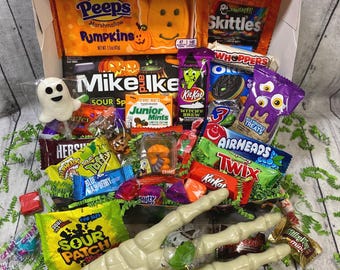 May include: A Halloween-themed cardboard box filled with assorted candies including Peeps Pumpkins, Skittles, Mike and Ike, KitKat, Whoppers, Airheads, Twix, and Sour Patch Kids. A large plastic skeleton hand rests in front of the box with loose candy.