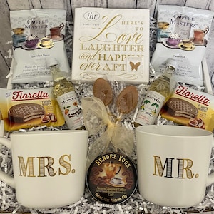 May include: A gift basket with a white background and a variety of coffee-themed items, including two mugs, one with "MRS." and the other with "MR." printed in gold, a small jar of coffee, two packets of coffee, two small bottles of flavoured syrup, two chocolate wafers, and a sign that reads "Here's to Love, Laughter and Happy Ever After".