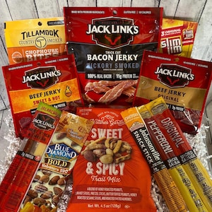Peut inclure: Un panier cadeau rempli de diverses collations sal&eacute;es. L'assortiment comprend du jerky, du m&eacute;lange montagnard, des b&acirc;tonnets de collation et des Slim Jim. Des marques comme Jack Link's, Tillamook et Blue Diamond sont visibles. L'emballage pr&eacute;sente des couleurs vives et des descriptions de produits.
