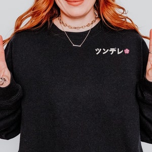 May include: Black sweatshirt with the Japanese word "Tsundere" embroidered on the chest, along with a small pink flower. The person is wearing a necklace and rings, and is making a peace sign with their fingers.