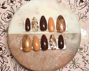 Peut inclure: Ensemble de dix faux ongles avec un motif brun, beige et or. Les ongles sont disposés sur une surface en marbre blanc.