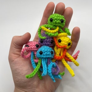 May include: A group of six crocheted jellyfish in various colors, including pink, purple, blue, green, and yellow. The jellyfish are arranged in a pile, with the green jellyfish on top.