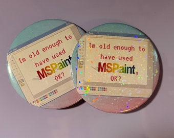 May include: Two round buttons with a glittery, iridescent finish. Each button features a simulated computer screen with the text "Im old enough to have used MSPaint, OK?" in a pixelated font.