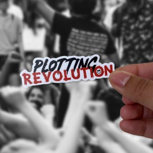 Könnte beinhalten: Ein weißer Aufkleber mit schwarzem und rotem Text, der "Plotting Revolution" mit einem Smiley-Symbol lautet. Der Aufkleber wird von einer Hand vor einem verschwommenen Hintergrund von Menschen gehalten.