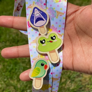 Puede incluir: Tres charms de madera en forma de paleta de helado con un diseño de tiburón, rana y pájaro. Los charms están unidos a un cordón blanco y rosa con lunares.