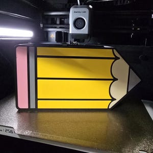 May include: A 3D printer is creating a pencil-shaped object. The design features a yellow body, a pink eraser, and a grey band. The printer is labelled "Bambu Lab". The pencil is a fun, educational design.
