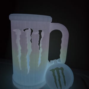 May include: A glowing, white 3D-printed beer mug with a handle and a matching lid. The mug features the Monster Energy logo and claw marks. The lid is round and also has the Monster Energy logo.