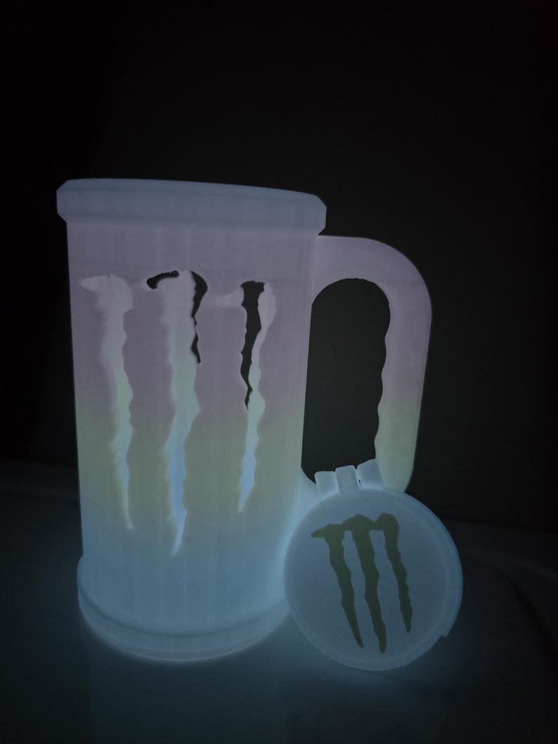 May include: A glowing, white 3D-printed beer mug with a handle and a matching lid. The mug features the Monster Energy logo and claw marks. The lid is round and also has the Monster Energy logo.