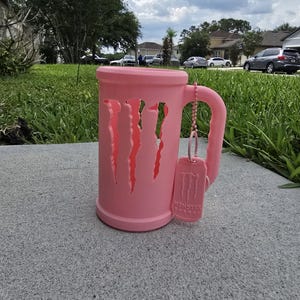 May include: A pink mug with cut-out claw marks, a handle, and a matching tag with the "Monster Energy" logo. The mug is sitting on a gray surface, with a green lawn and houses in the background.