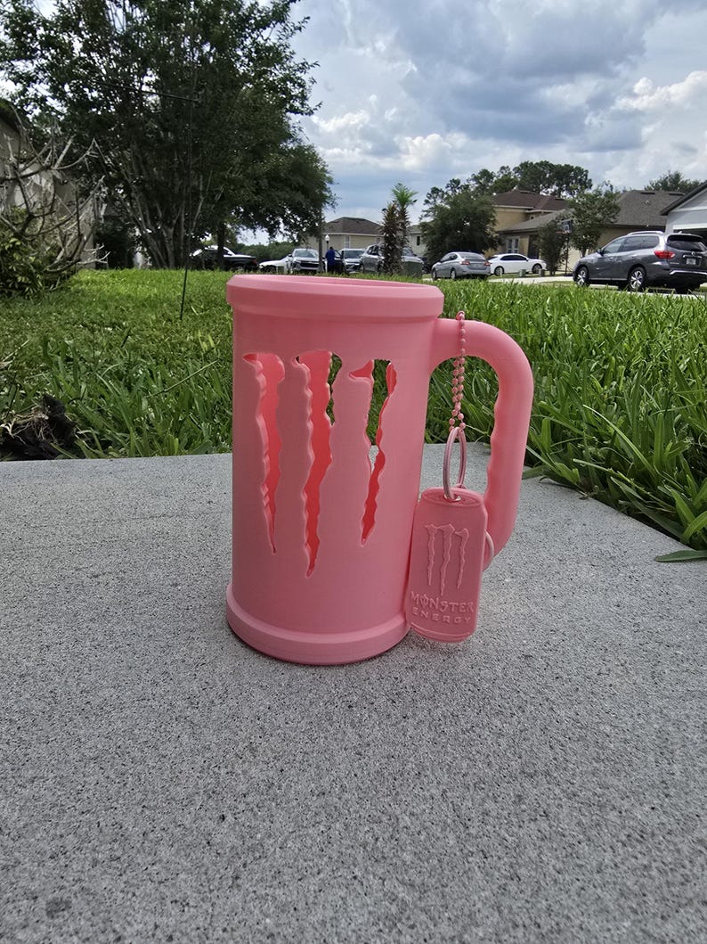 May include: A pink mug with cut-out claw marks, a handle, and a matching tag with the "Monster Energy" logo. The mug is sitting on a gray surface, with a green lawn and houses in the background.