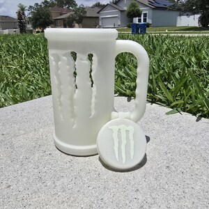 May include: A white, frosted glass beer mug with a handle and a matching coaster. The mug features a design of claw marks cut into the side. The coaster has the Monster Energy logo in green. The mug and coaster are sitting on a concrete surface.