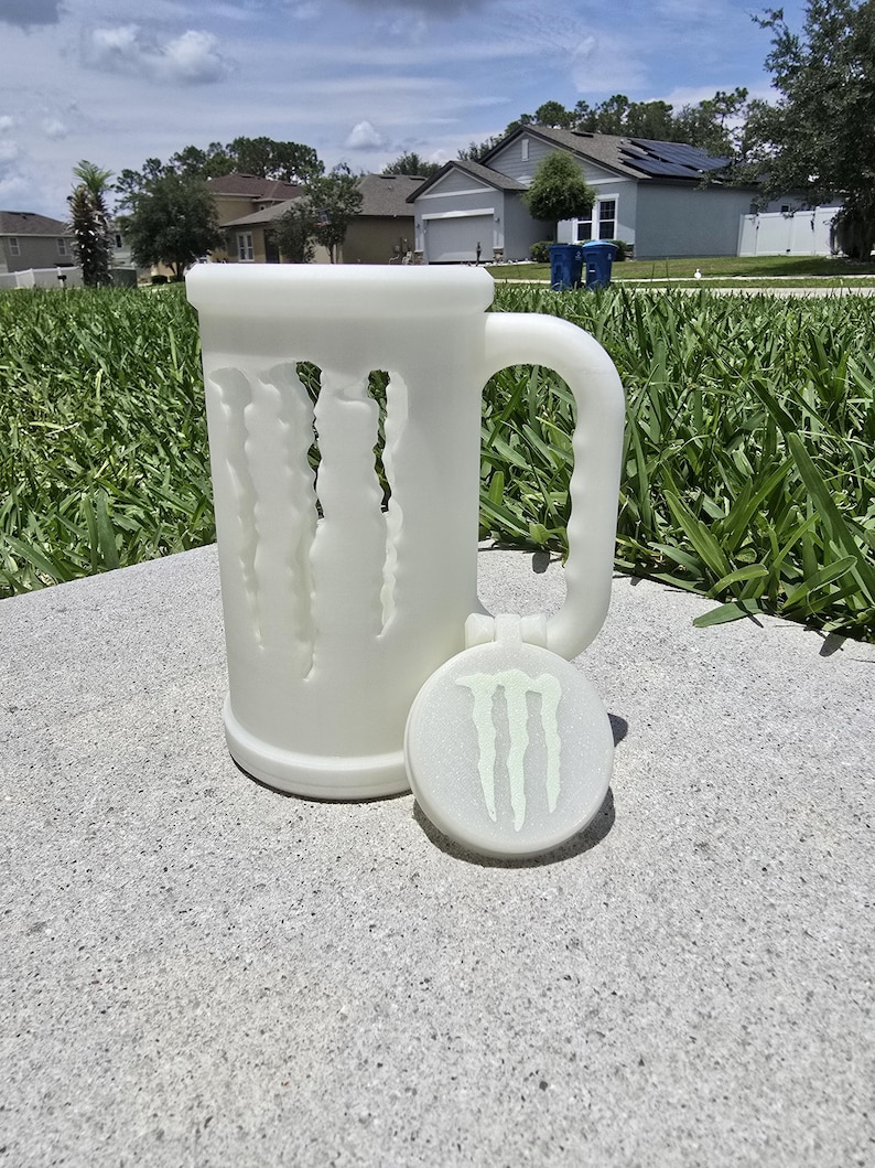 May include: A white, frosted glass beer mug with a handle and a matching coaster. The mug features a design of claw marks cut into the side. The coaster has the Monster Energy logo in green. The mug and coaster are sitting on a concrete surface.
