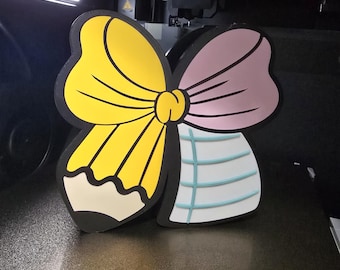 May include: A 3D-printed, multi-colored bow-shaped object. One side is yellow with a pencil design, the other is pink. The center is a knot, and the bottom section is white with blue lines.