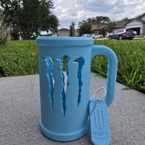 May include: A light blue mug with a handle and a tag. The mug has claw-like cutouts on the side. The tag reads "Monster Energy". The mug is sitting on a gray surface.