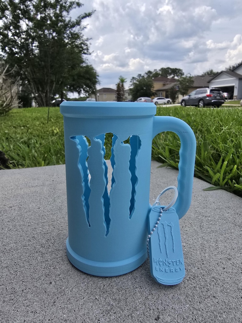 May include: A light blue mug with a handle and a tag. The mug has claw-like cutouts on the side. The tag reads "Monster Energy". The mug is sitting on a gray surface.