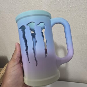 May include: A large, ombre-colored mug with a handle. The mug transitions from light blue to purple and green. Three claw-shaped cutouts are visible on the side. The mug is held by a hand.