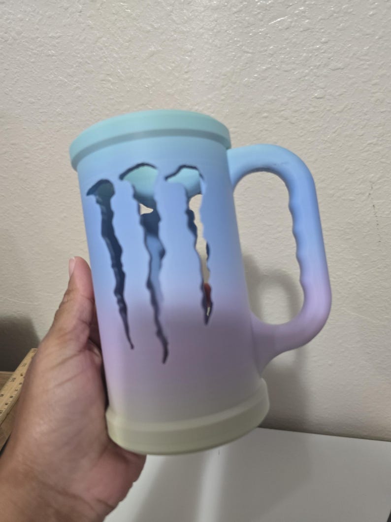 May include: A large, ombre-colored mug with a handle. The mug transitions from light blue to purple and green. Three claw-shaped cutouts are visible on the side. The mug is held by a hand.