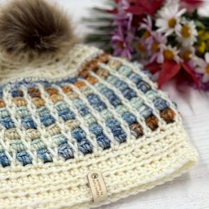 May include: Cream-colored crochet beanie with a brown faux fur pom-pom. The hat features a textured pattern with blue, orange, and tan accents. A small tag is attached to the brim. A bouquet of flowers is in the background.