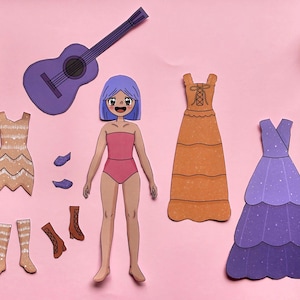 Puede incluir: Un set de muñeca de papel que incluye un personaje con cabello morado en traje de baño rosa, una guitarra acústica morada, un vestido corto beige con patrones de chevrón, un vestido largo naranja, un vestido morado escalonado y dos pares de zapatos.