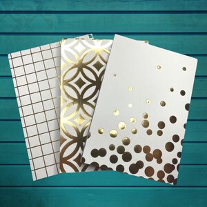 Może przedstawiać: Trzy białe zeszyty z wzorami z złotej folii. Zeszyty mają geometryczne wzory, kropki i koła.