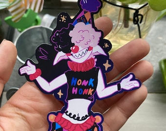 May include: A sticker of a cartoon character with pink fur, black hair, and a red and white striped hat. The character is wearing a black shirt with the words "Honk Honk" printed on it. The character is also wearing a colorful skirt with a black and white checkered pattern. The sticker has a glittery finish.